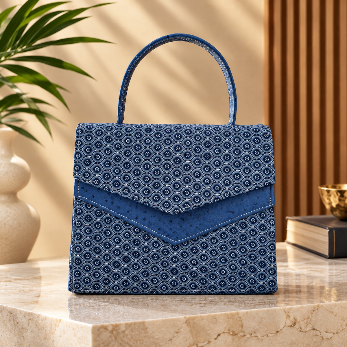 A structured blue handbag with a repeating geometric pattern and a top handle, displayed on a marble surface in a styled interior setting with a plant and books in the background.
