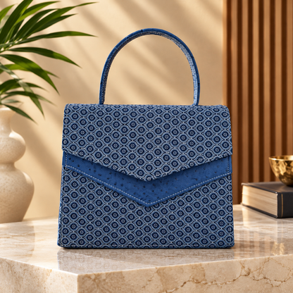 A structured blue handbag with a repeating geometric pattern and a top handle, displayed on a marble surface in a styled interior setting with a plant and books in the background.